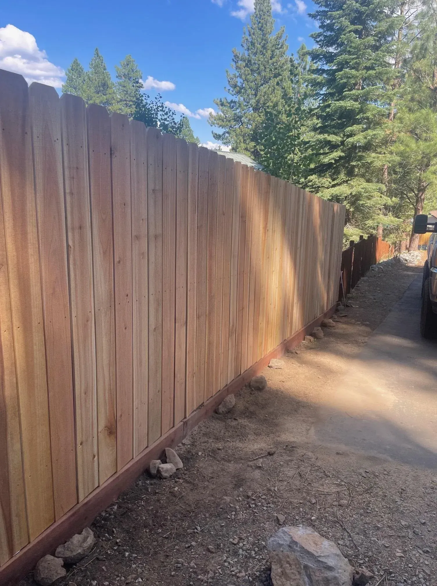Wooden fence next to a dirt driveway, with trees in the background under a blue sky.