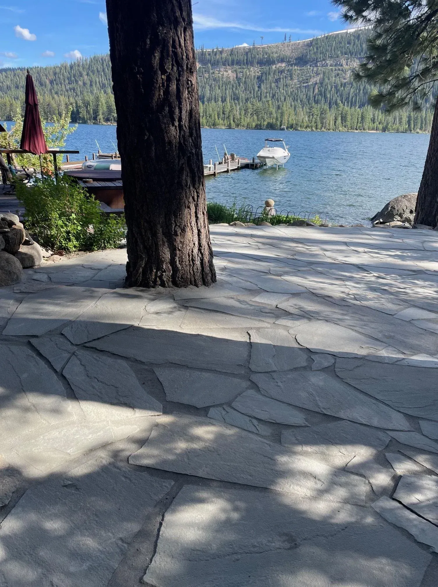 Stone patio leading to a lake with a boat, trees, and dock under a sunny sky.