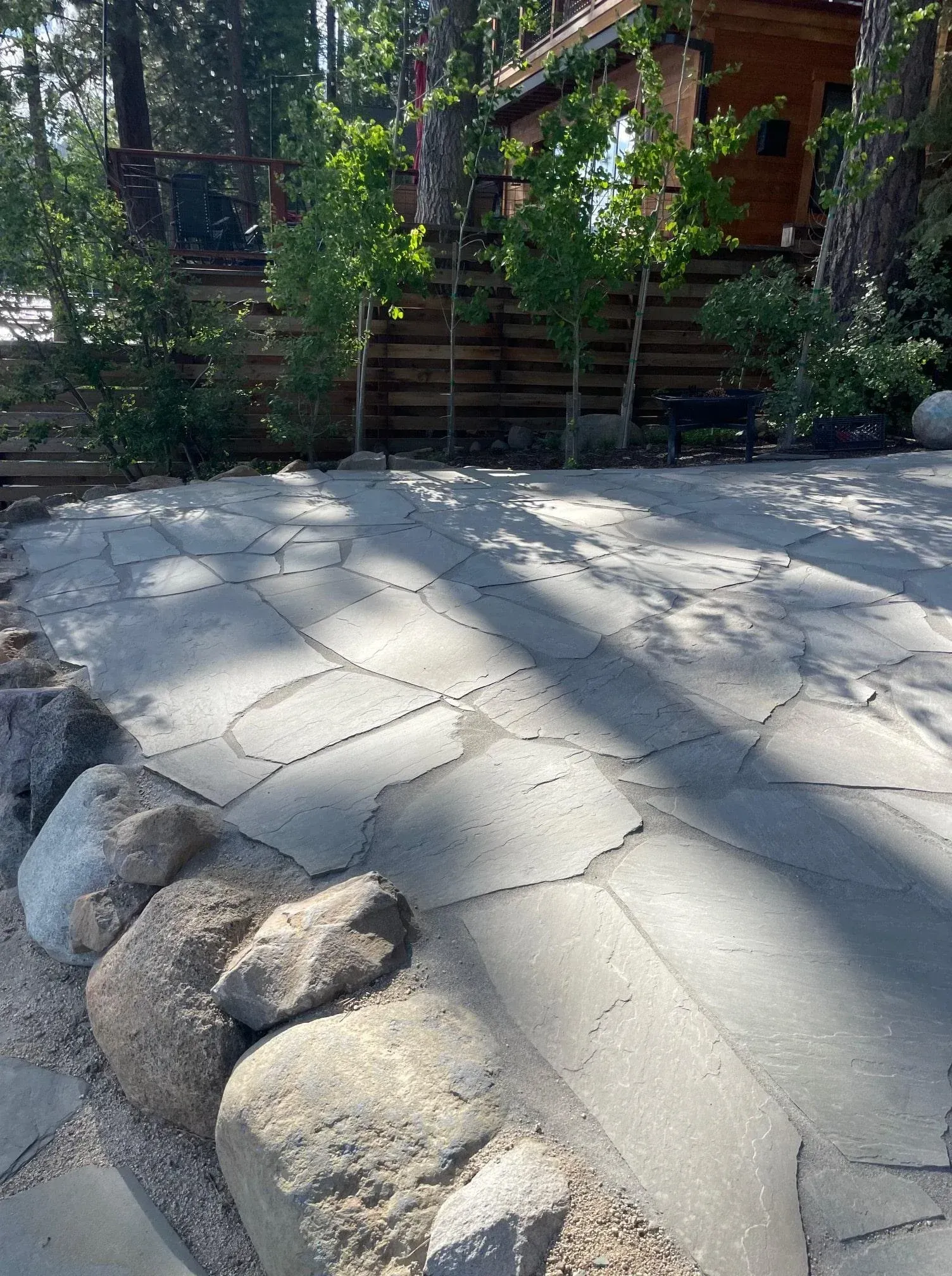 A paved area with large rocks bordering the edge. Young trees and a wooden fence are in the background.