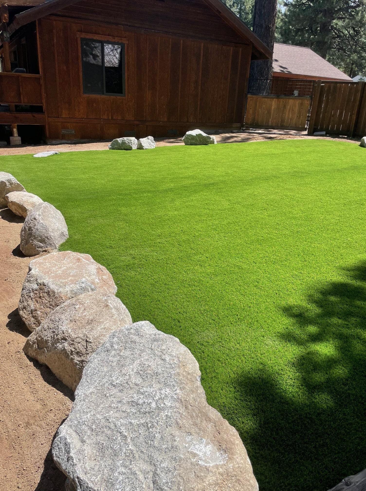 Lush green artificial turf in backyard surrounded by rocks and a wooden cabin.