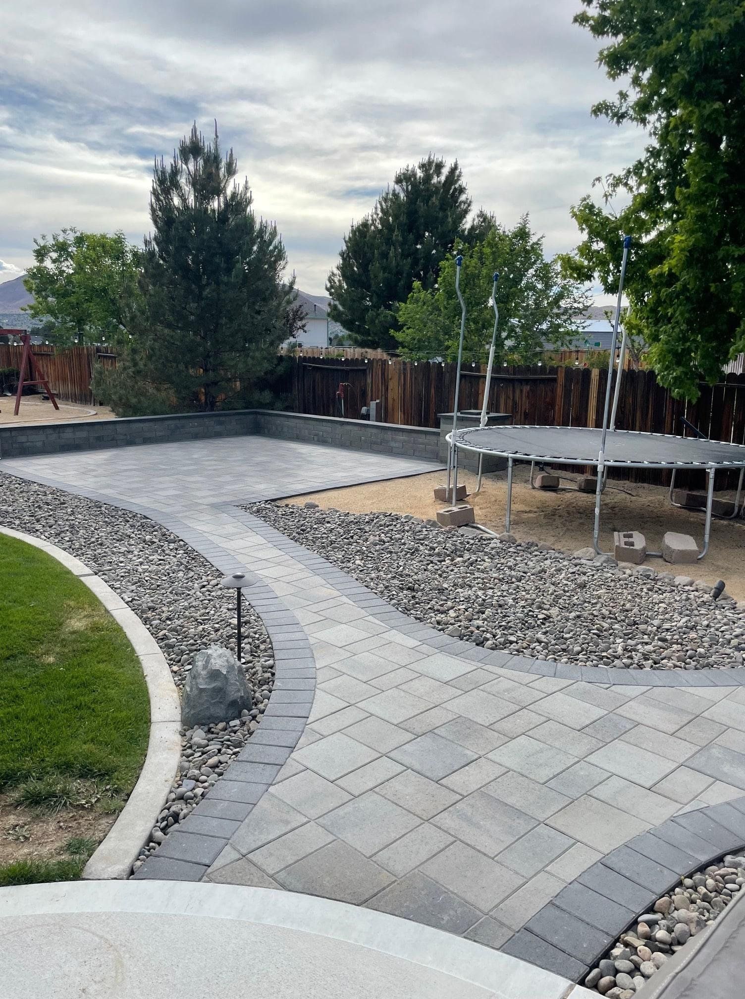 Backyard with stone patio, path, and rock landscaping; trampoline and trees in the background.