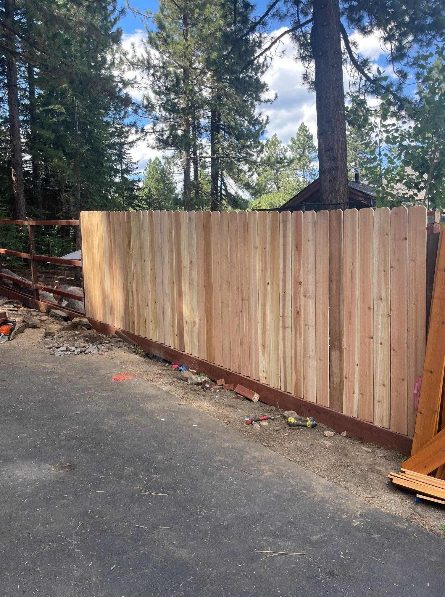 New wooden fence in front of a driveway, with trees and a house in the background.