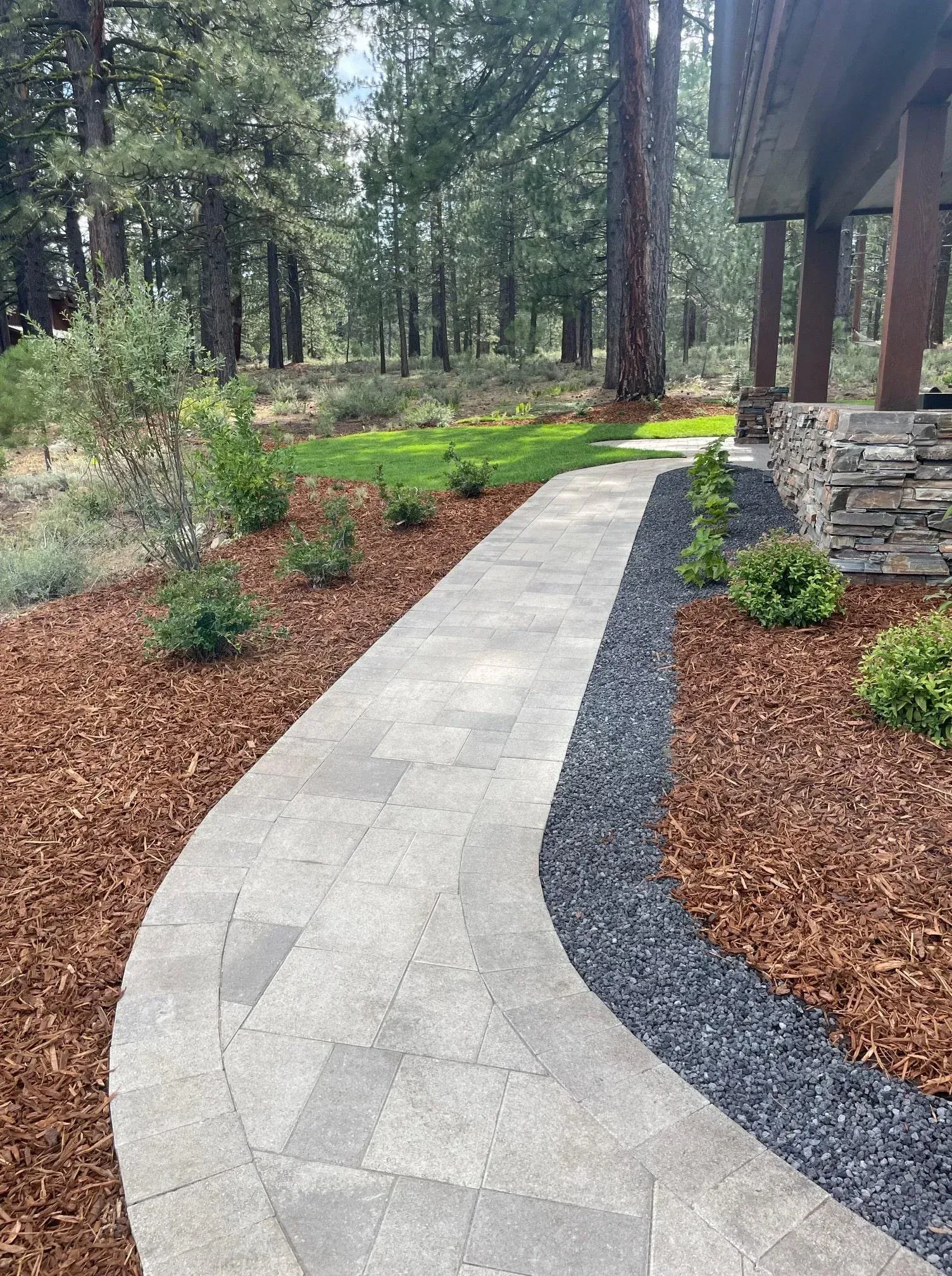 Stone pathway curves through a landscaped yard with mulch, grass, bushes, and a dark gravel border.