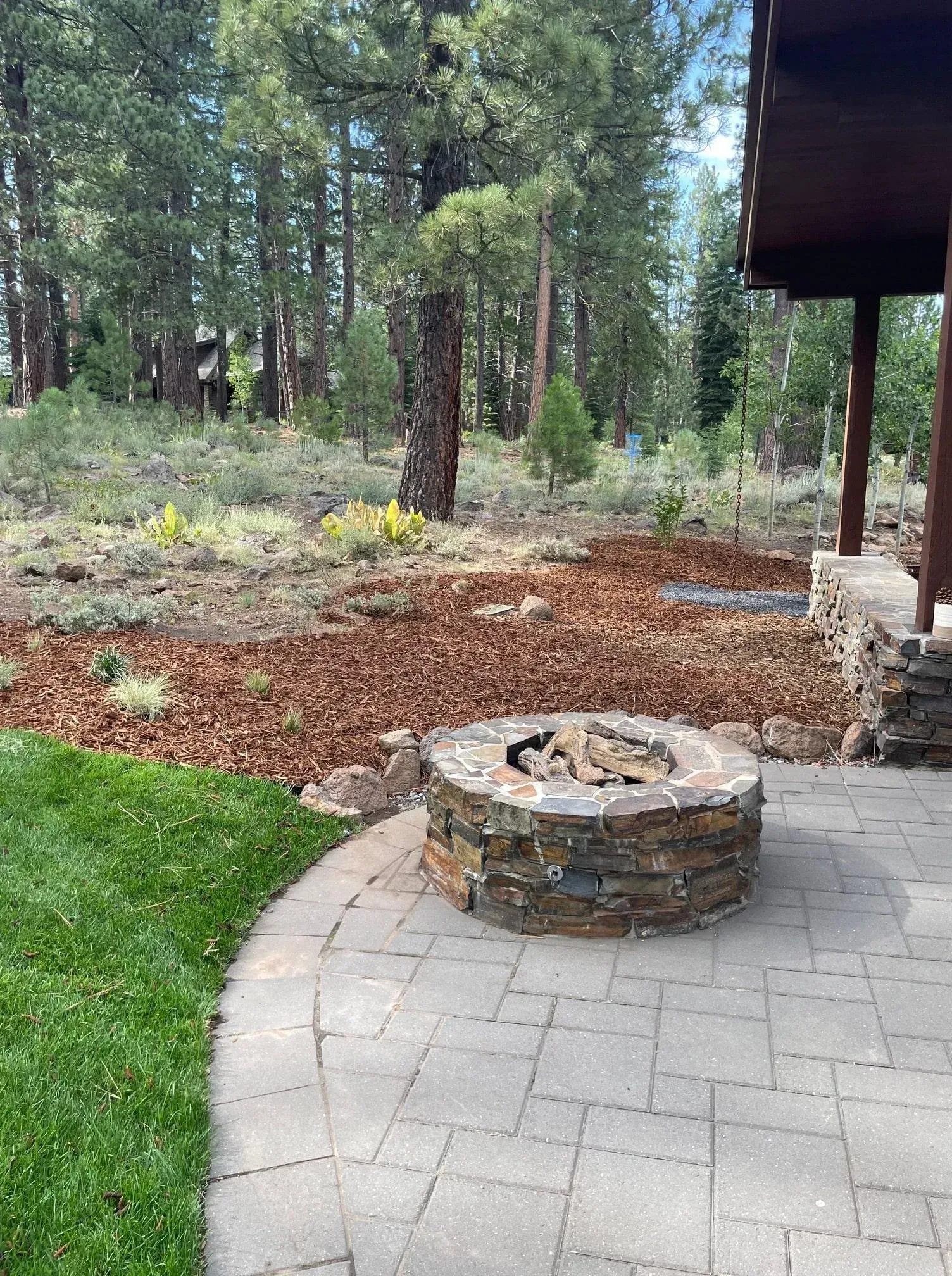 Stone fire pit on a patio, surrounded by mulch, grass, and trees.