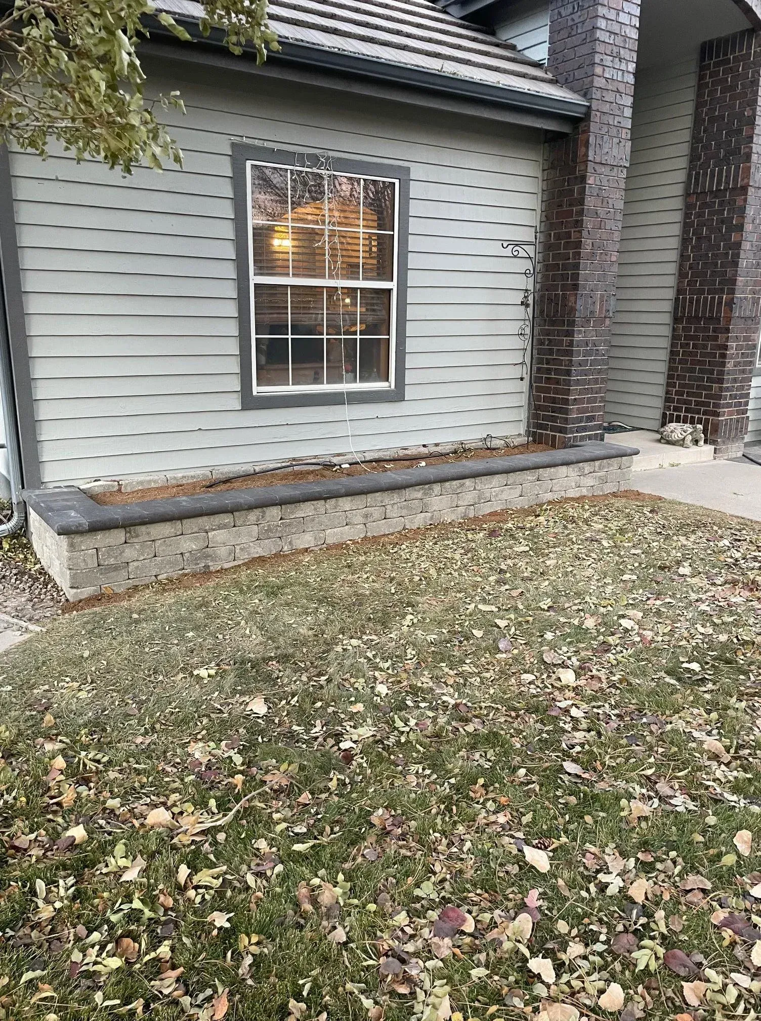 Low retaining wall in front of a house. Window above the wall, siding, and brown and green grass are visible.