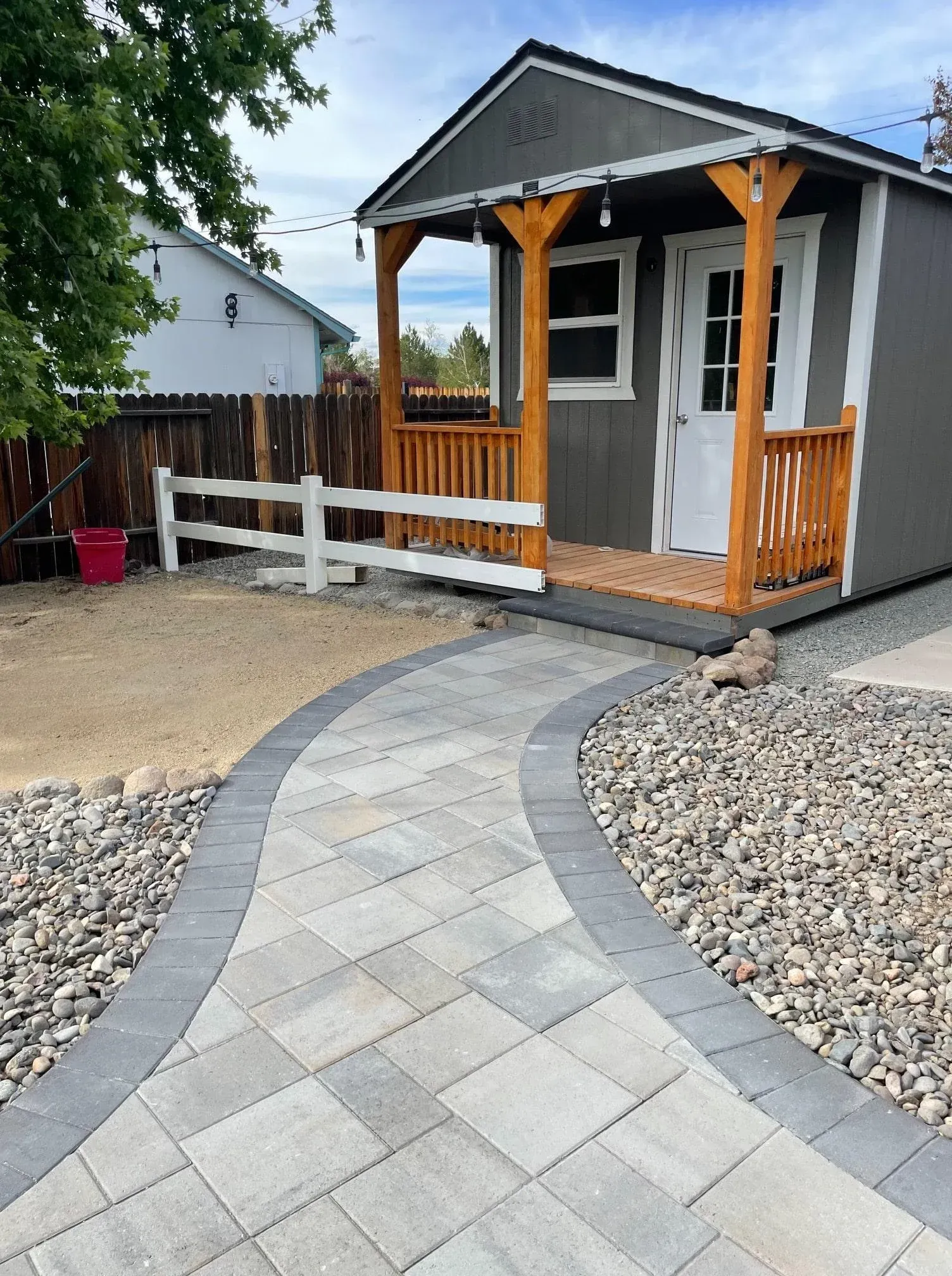 Pathway leading to a gray shed with wooden accents; a white fence is in the background.