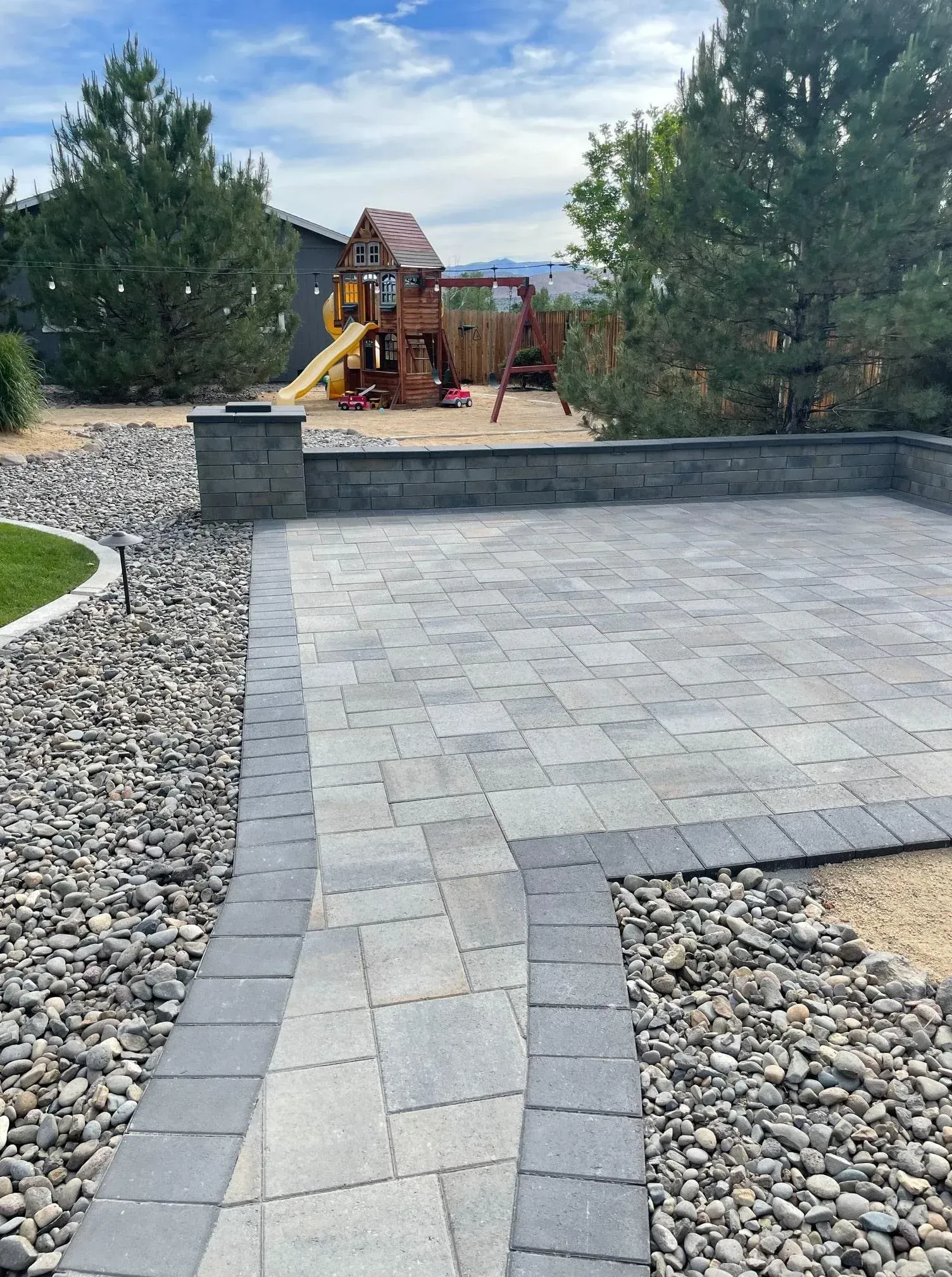 Paver patio and walkway with retaining wall, rock landscaping, and a playground in the background.
