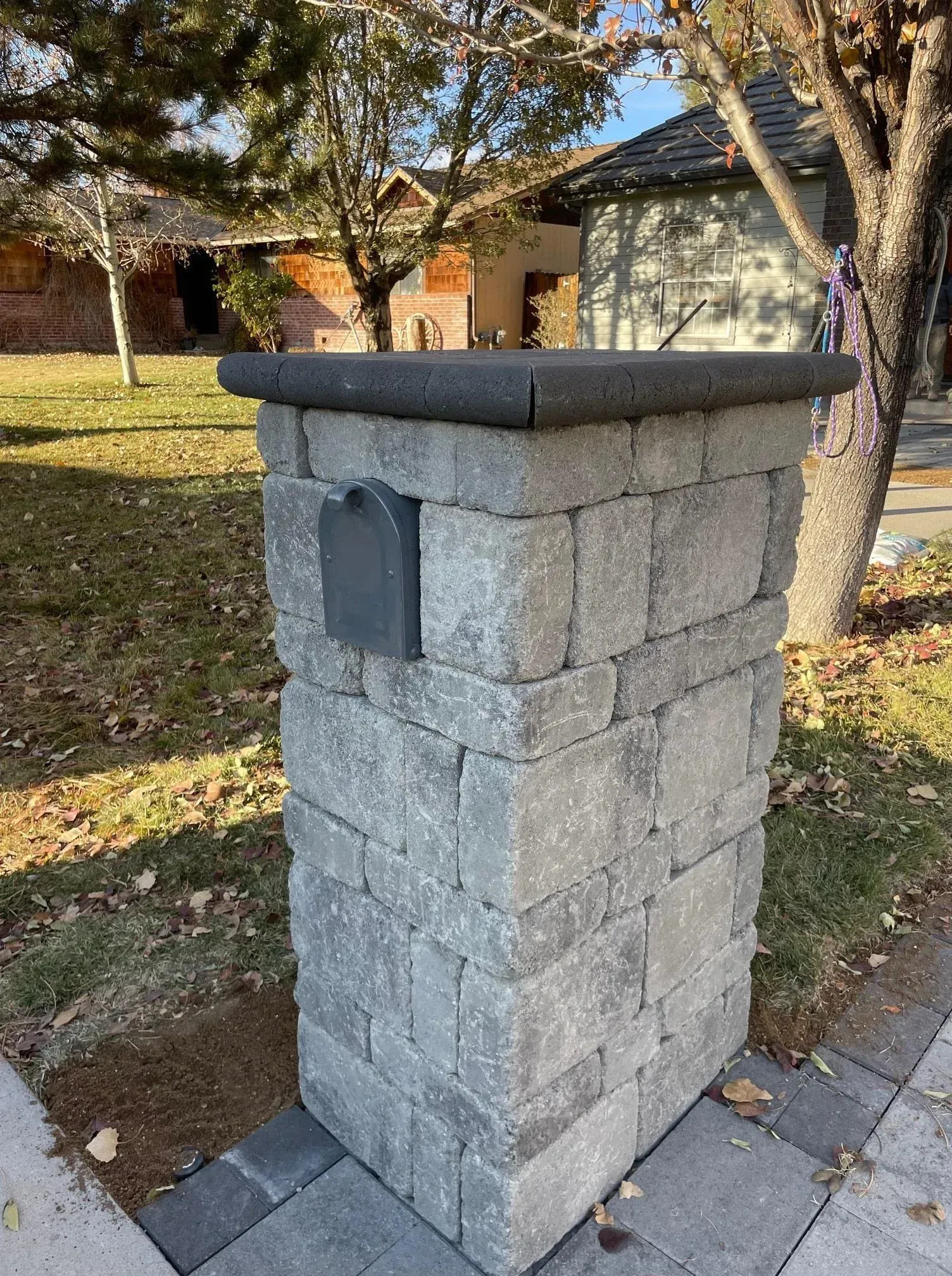 Gray brick mailbox post with a dark gray top, next to a sidewalk and lawn.