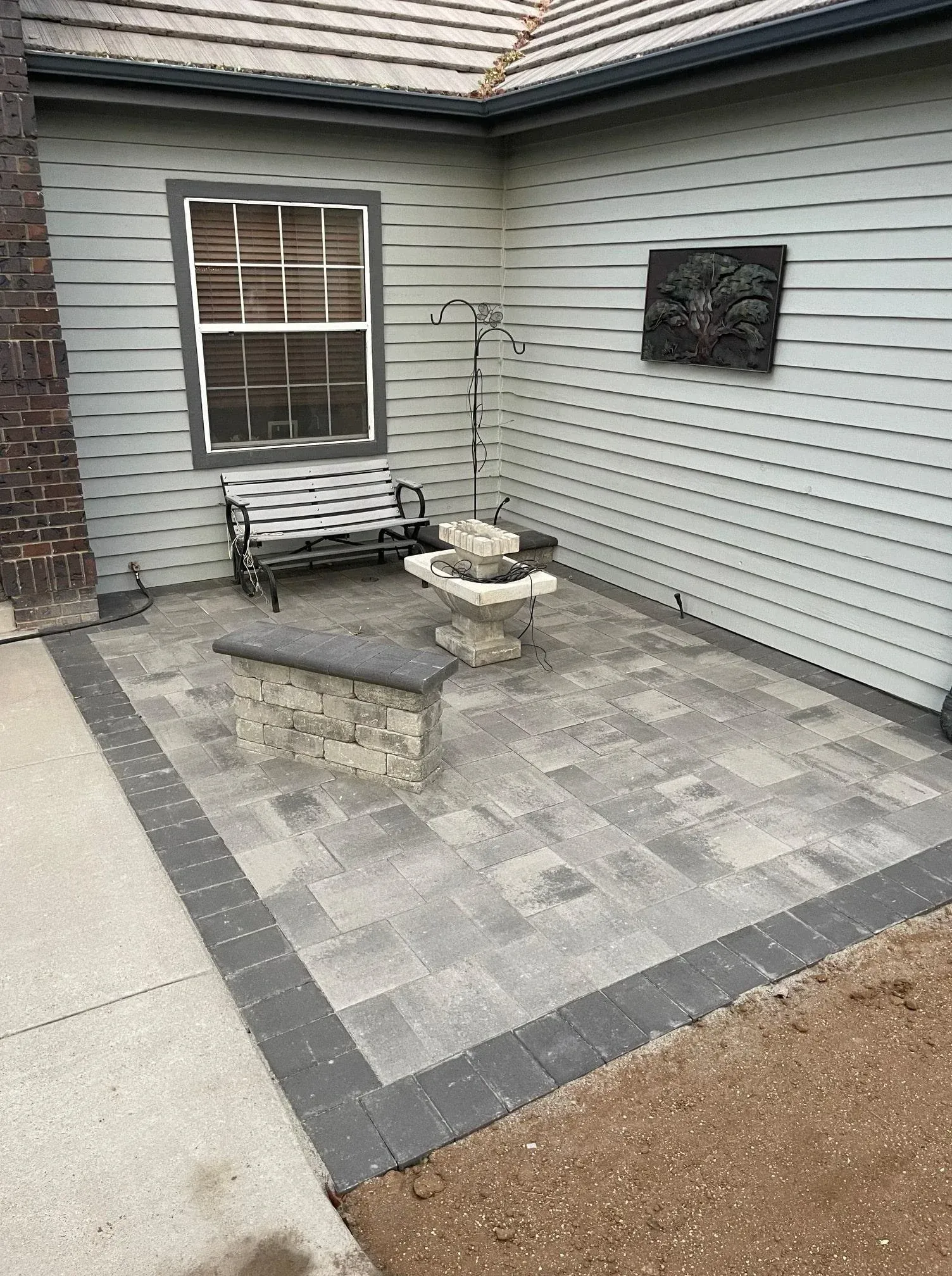 Patio with gray pavers, bench, decorative art, and a dark border, next to a house.