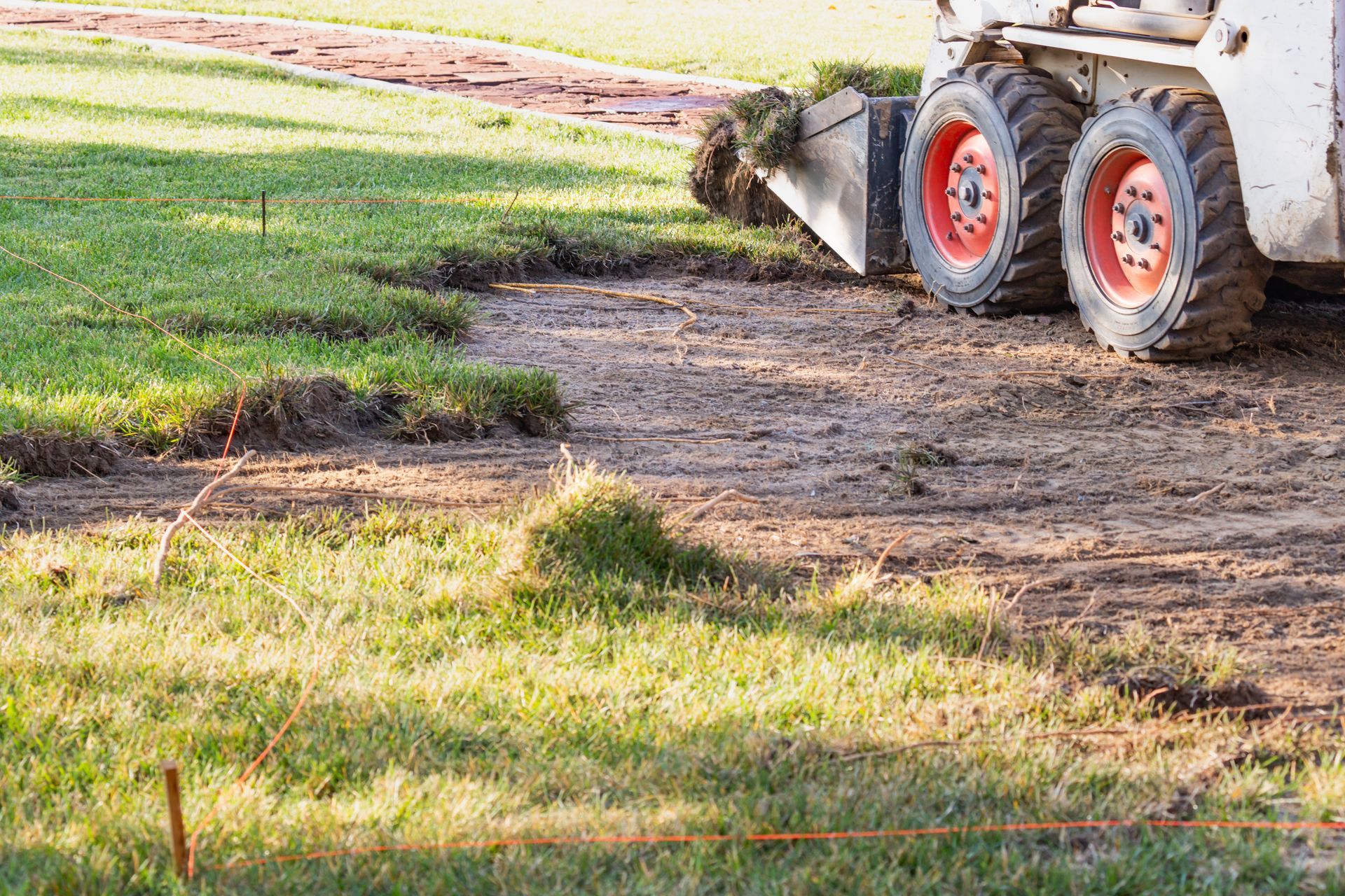 Skid steer removing sod from a muddy patch of grass. Red wheels, green and brown earth, sunny outdoors.