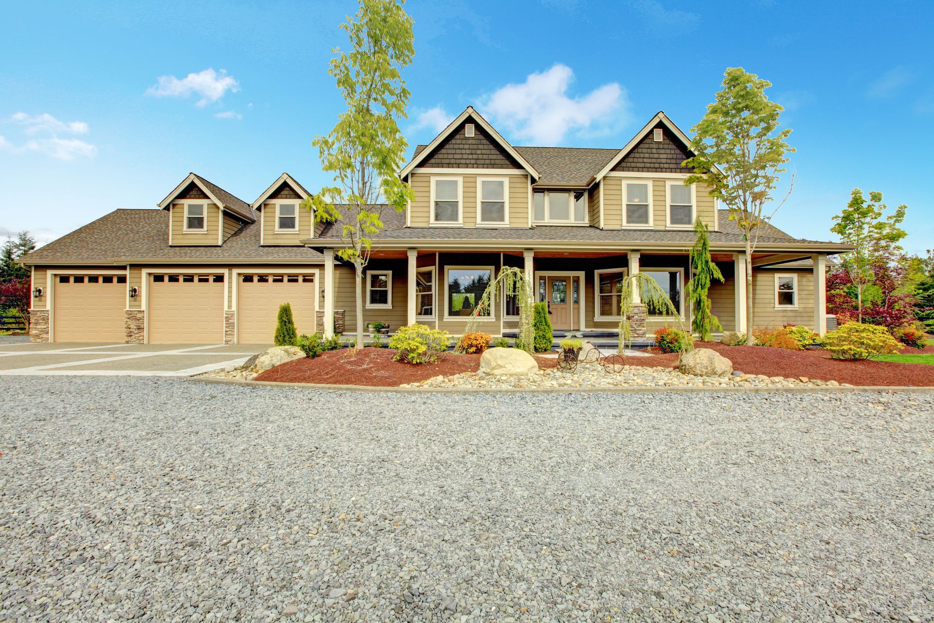 Two-story beige house with a gravel driveway, front porch, and three-car garage under a blue sky.