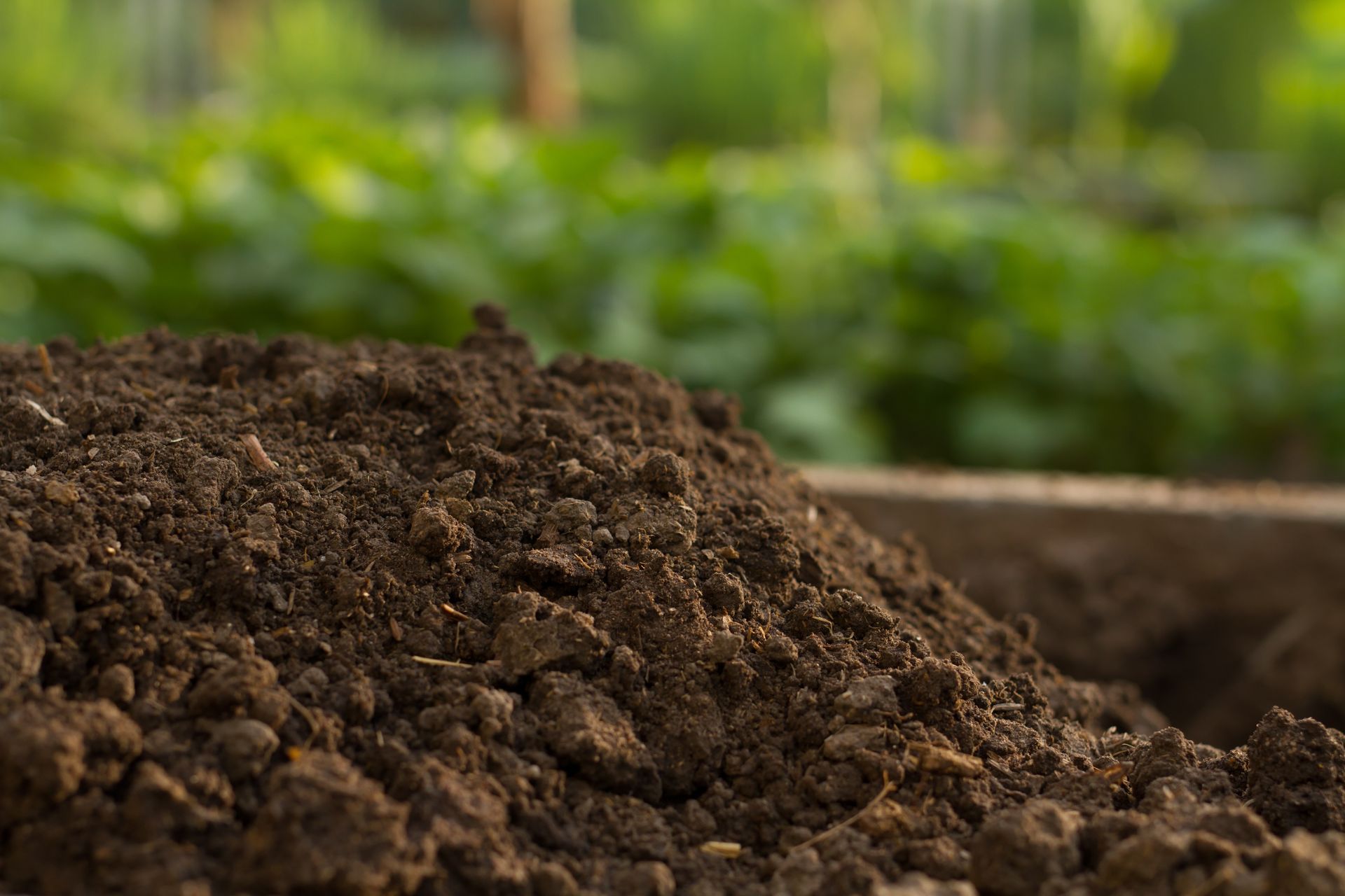 Pile of dark, rich soil in a wooden container, with blurred green foliage in the background.