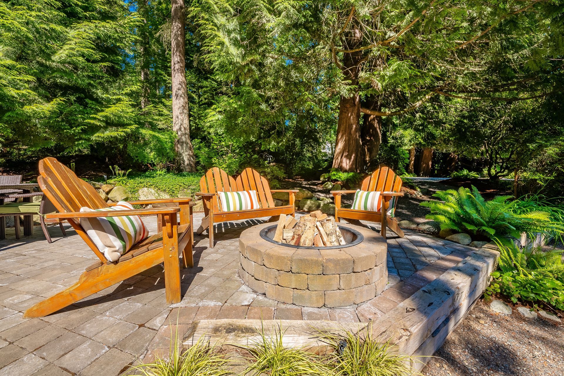 Four wooden Adirondack chairs around a brick fire pit on a patio, surrounded by trees.