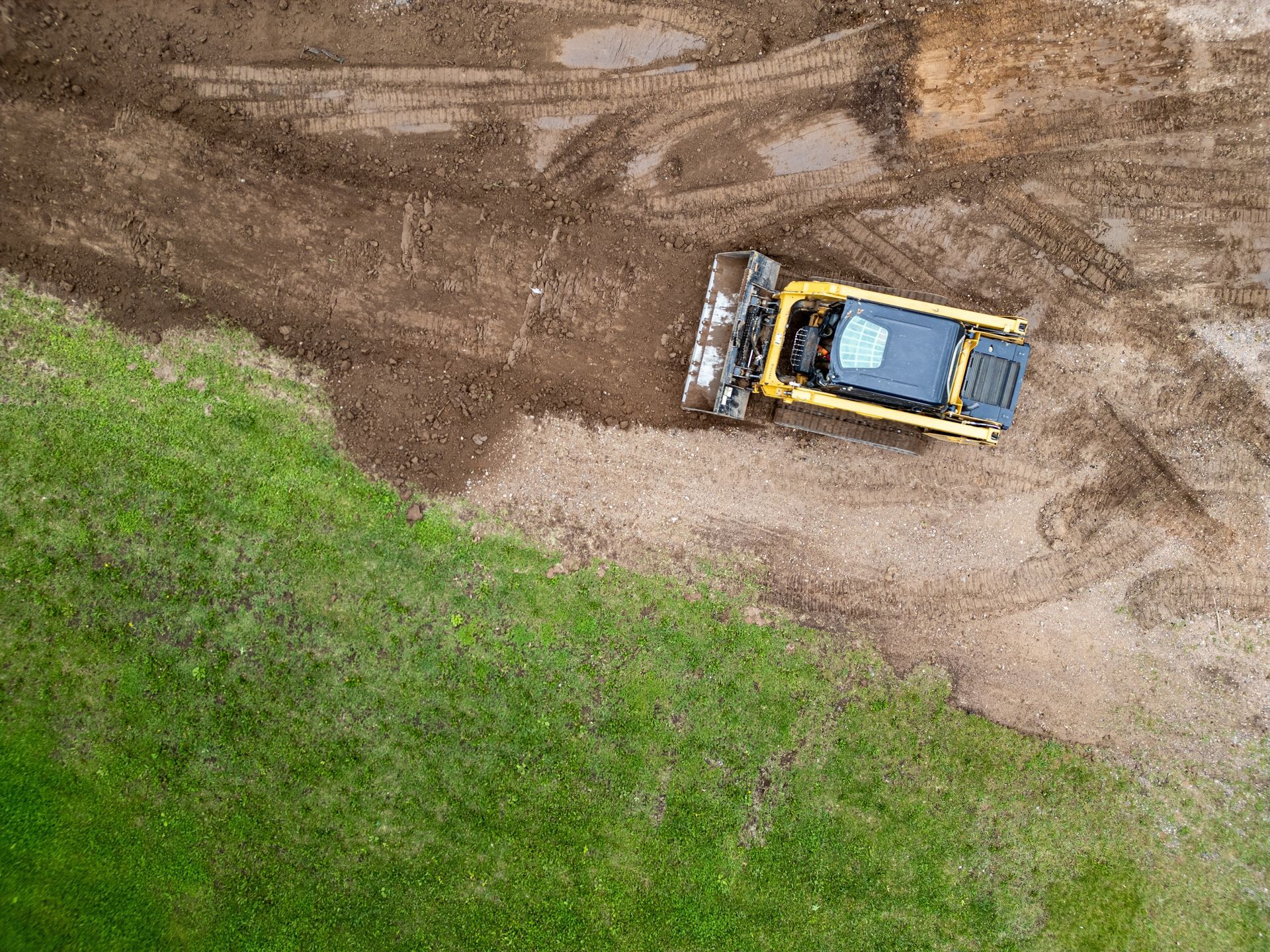 Overhead view: Yellow bulldozer on a dirt area next to a green grassy field.