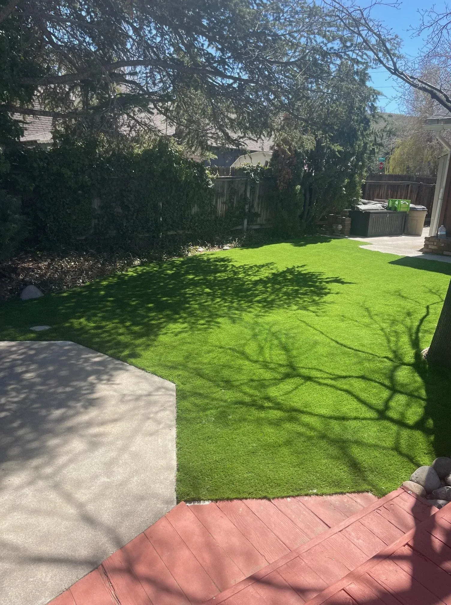 Lush green artificial lawn in a backyard. Concrete patio and red wood deck edge the grass.