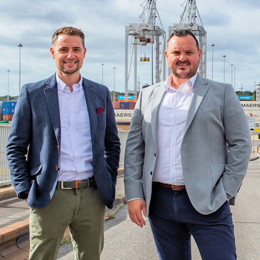 Two men in suits pose by a shipping port. One man wears a navy blazer, the other a grey blazer.