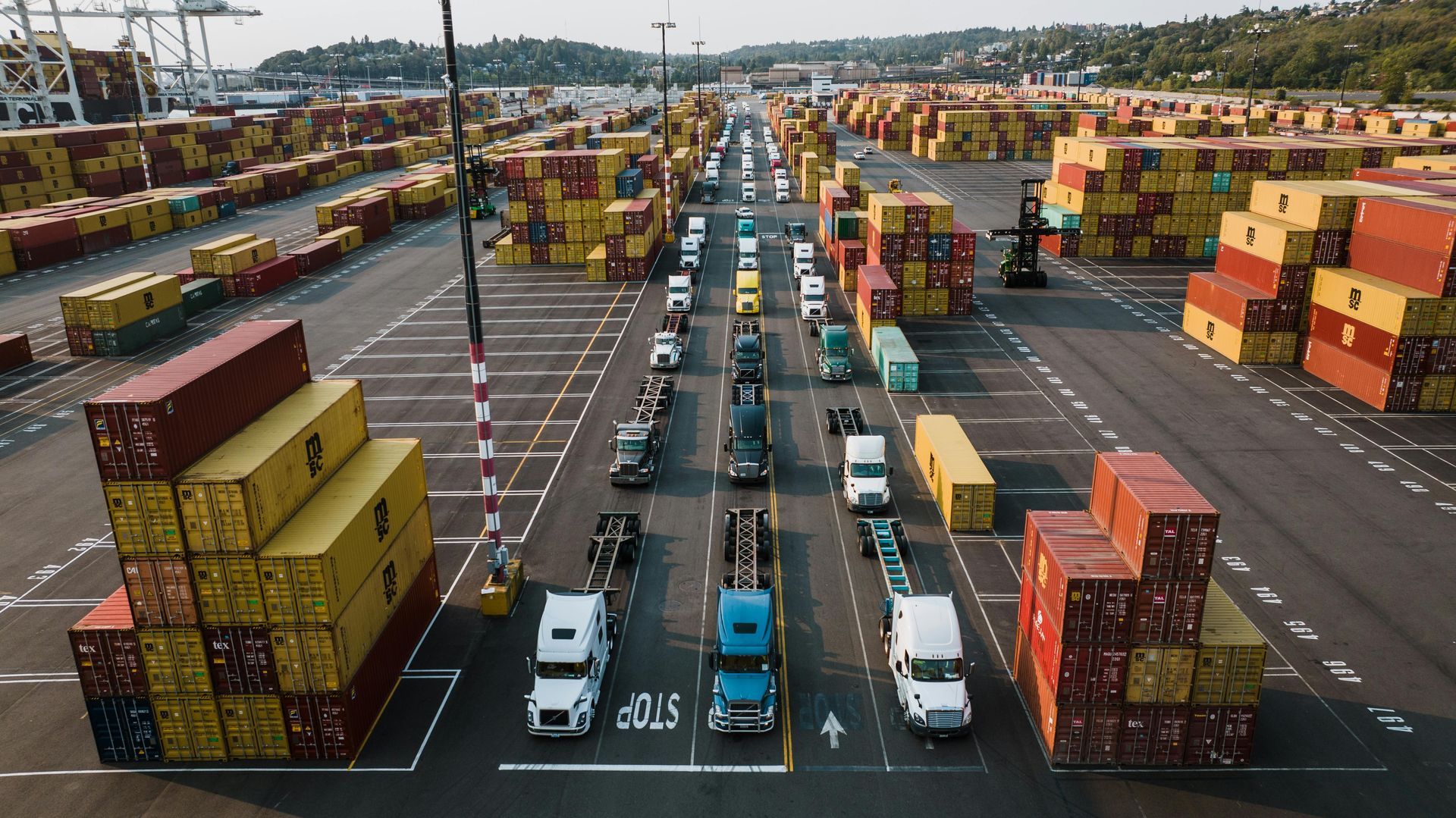 Cargo trucks line up in a port, surrounded by stacks of shipping containers in a wide outdoor shot.