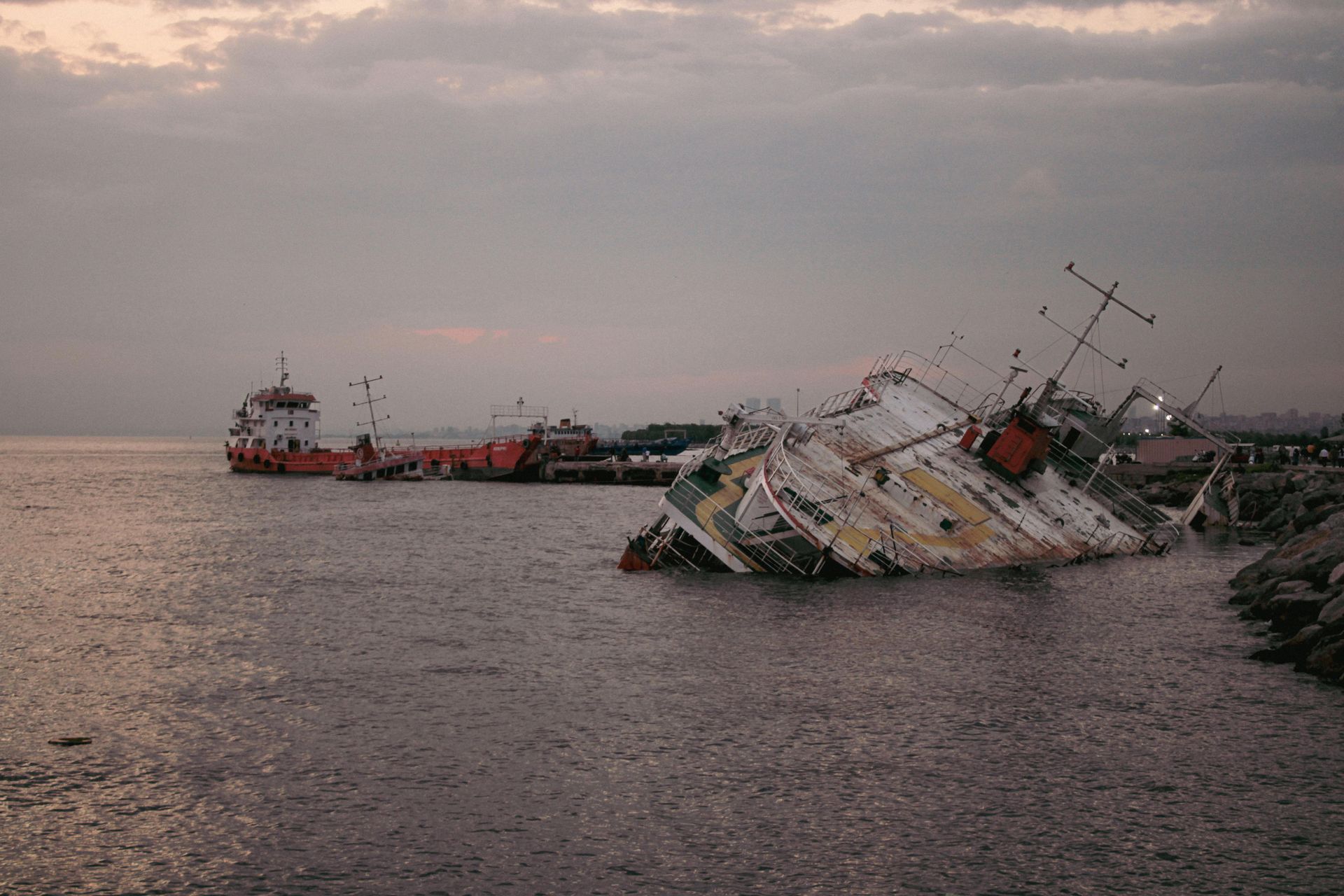 Sunken ship by a pier, with another ship in the background; grey water and cloudy sky.