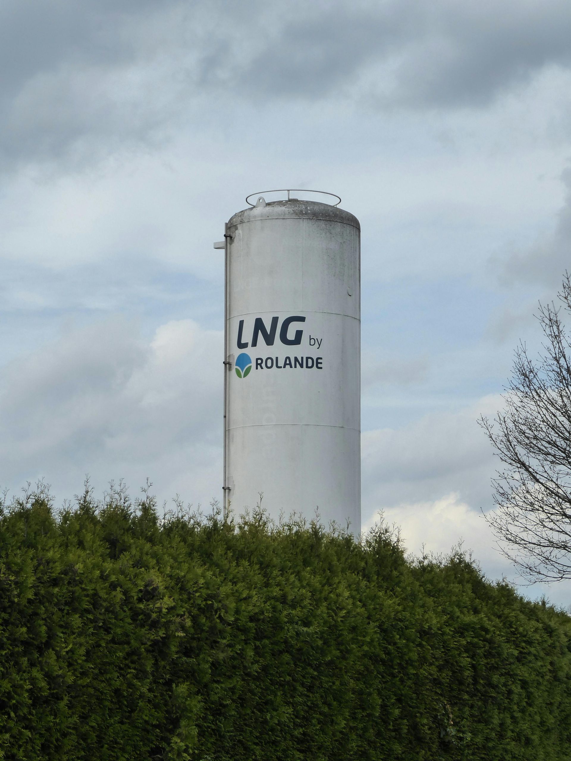 White LNG storage tank by Rolande, rising above green bushes under a cloudy sky.
