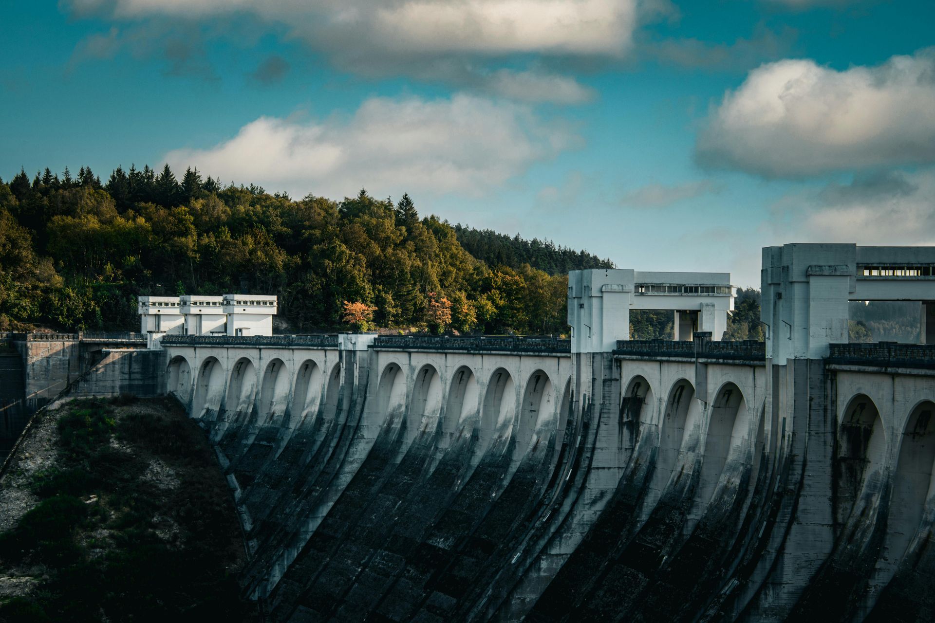 Concrete dam with white structures against a green forest and cloudy blue sky.
