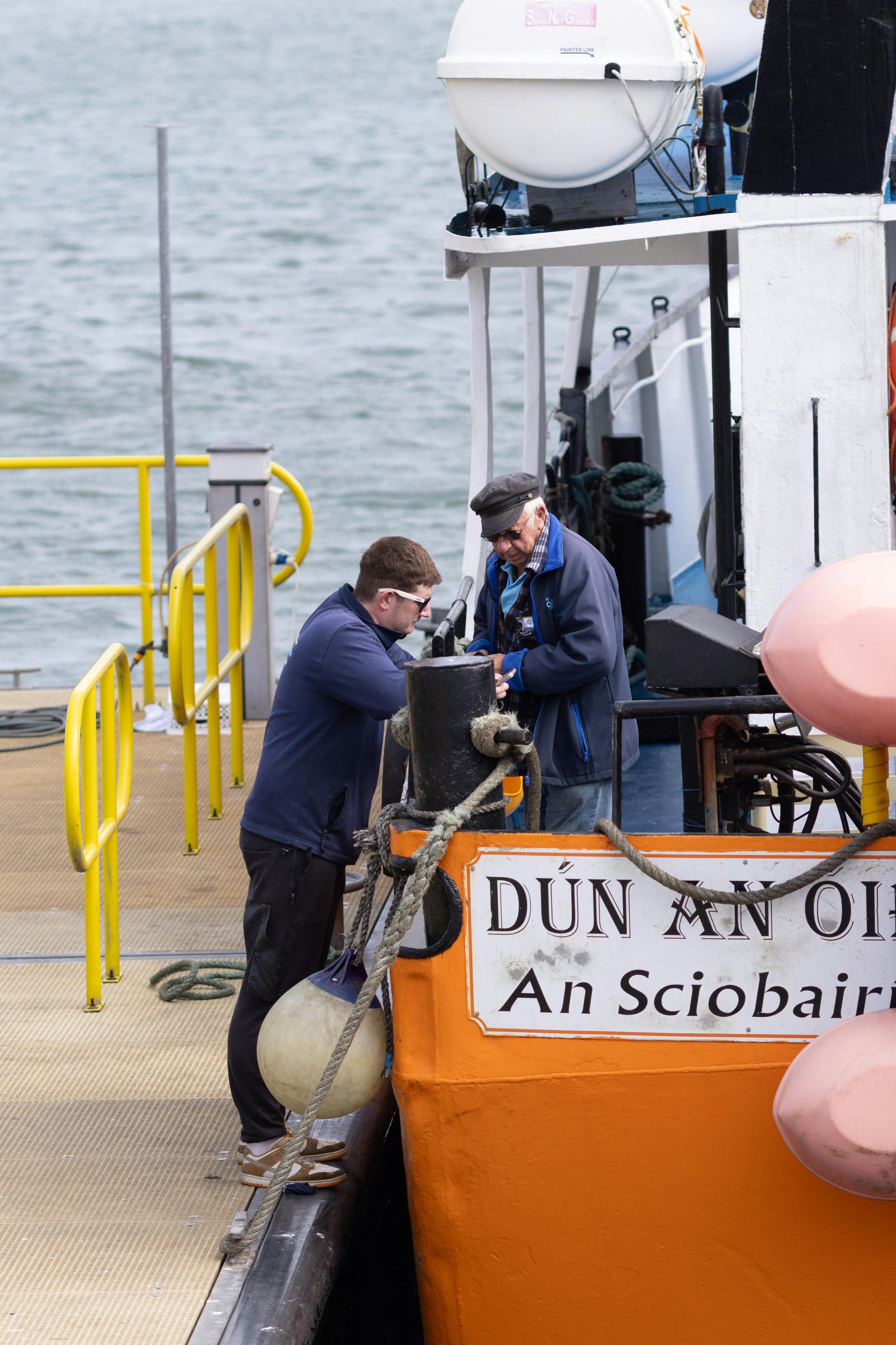Two men on a ferry; one in blue working on the boat, the other wearing a cap. Orange ferry side.