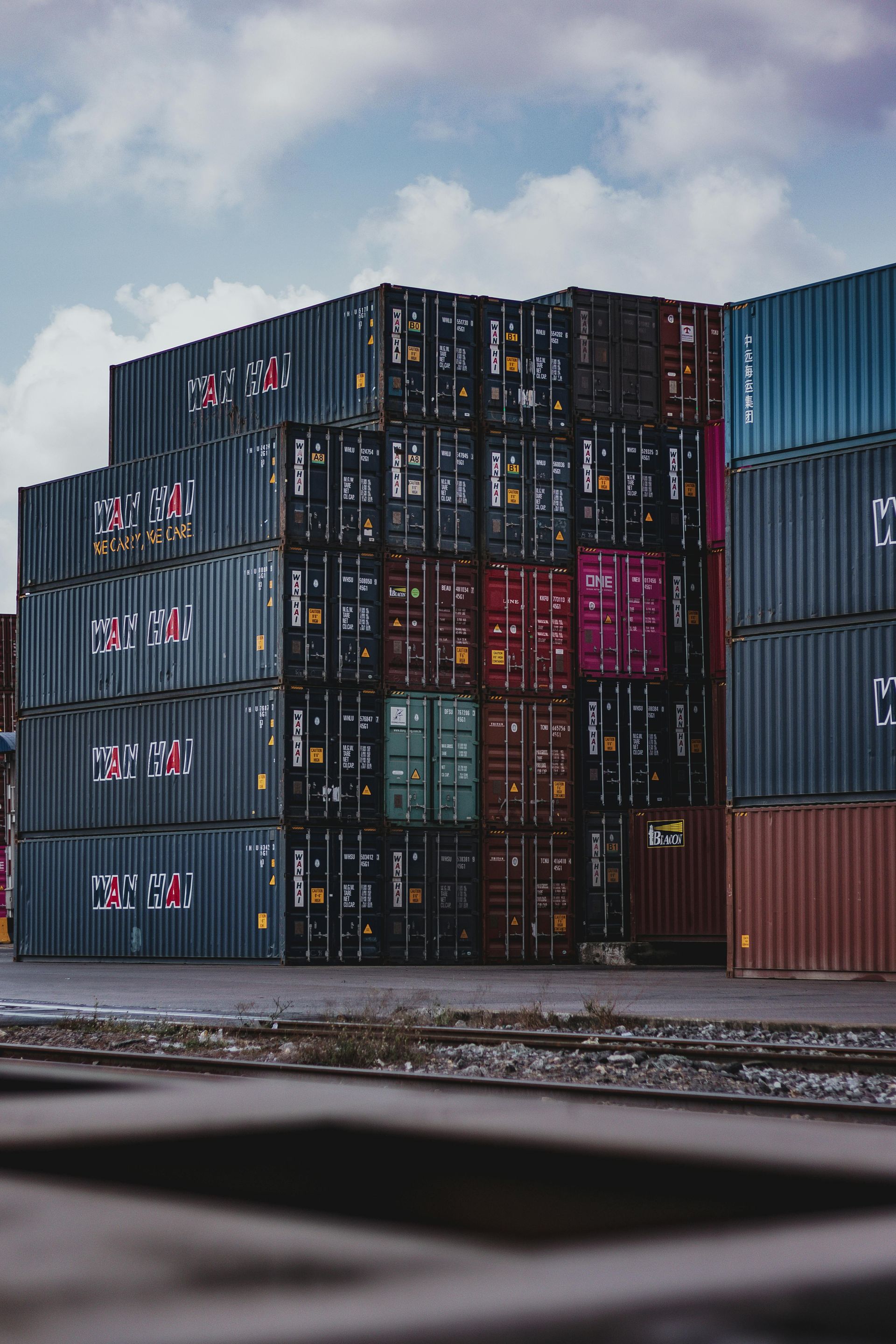 Stacks of colorful shipping containers at a port, under a cloudy sky.