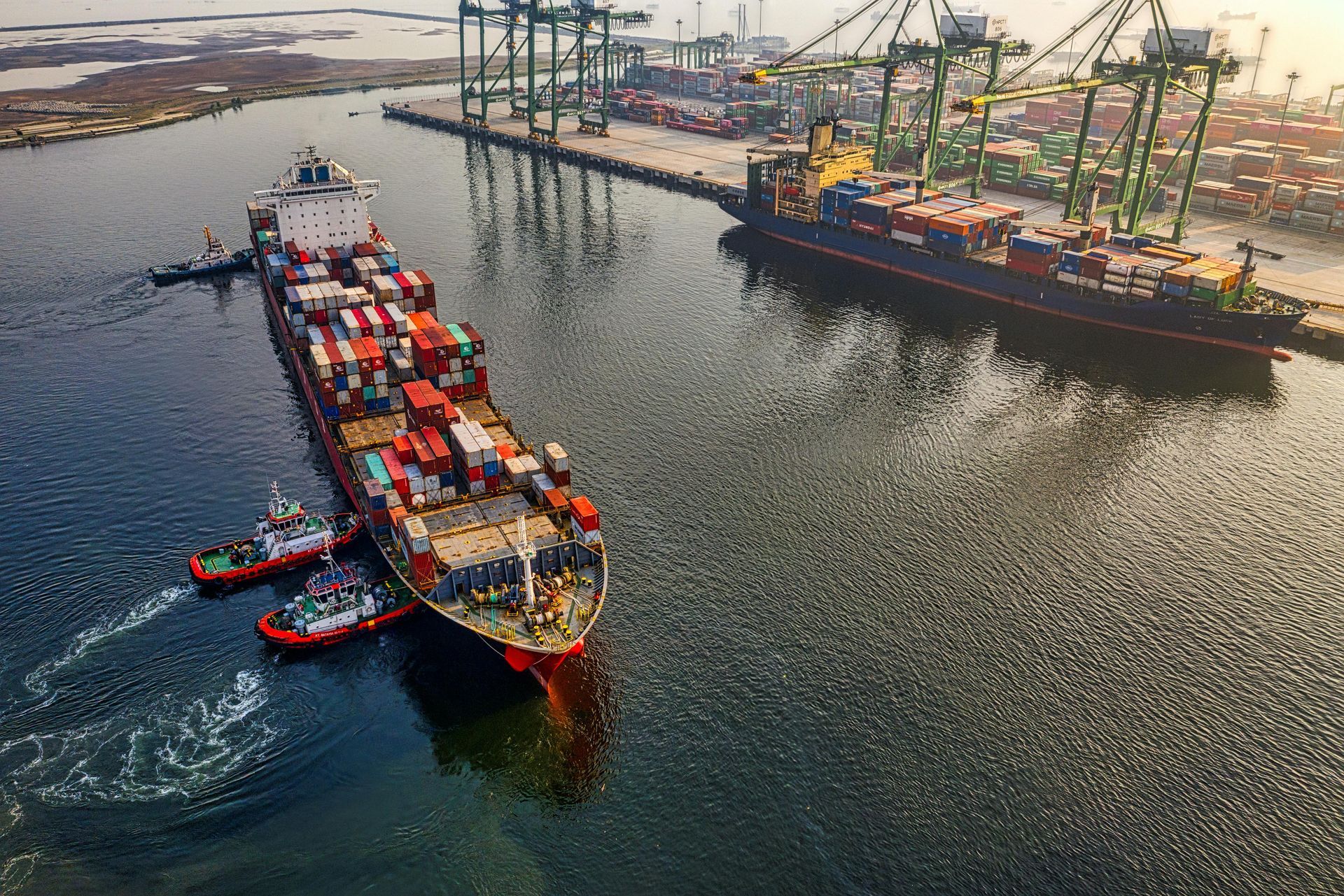 Large cargo ship being assisted into a port by tugboats; cranes, containers, and another ship are in view.