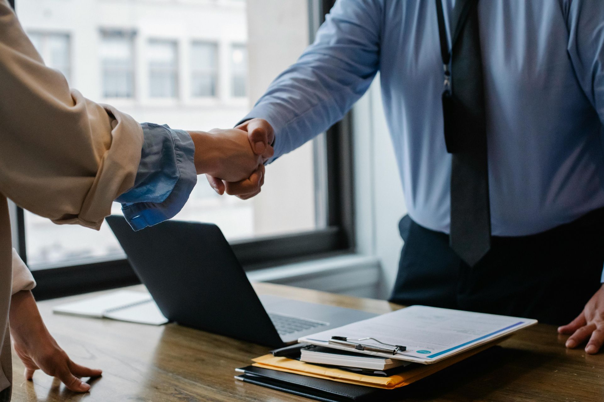 Two people shaking hands at a desk near a laptop and paperwork.