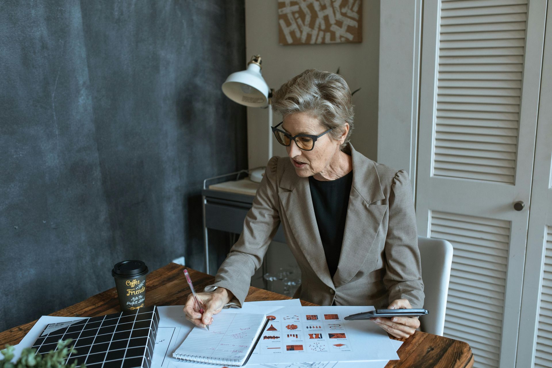 Woman in glasses and blazer working at a desk, writing on paper and looking at printouts.