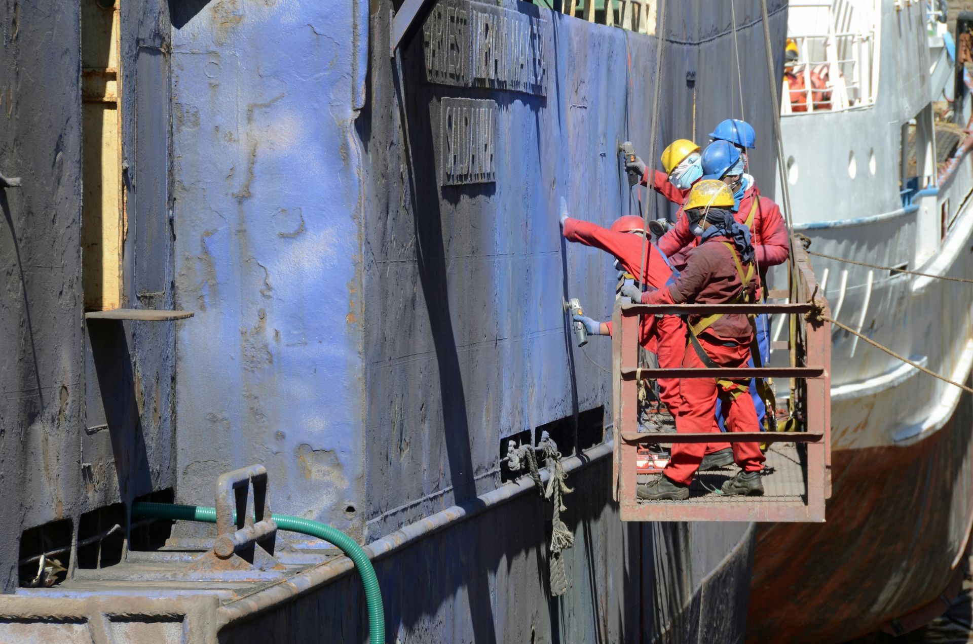 Workers in safety gear painting a large, blue metal structure on a ship.