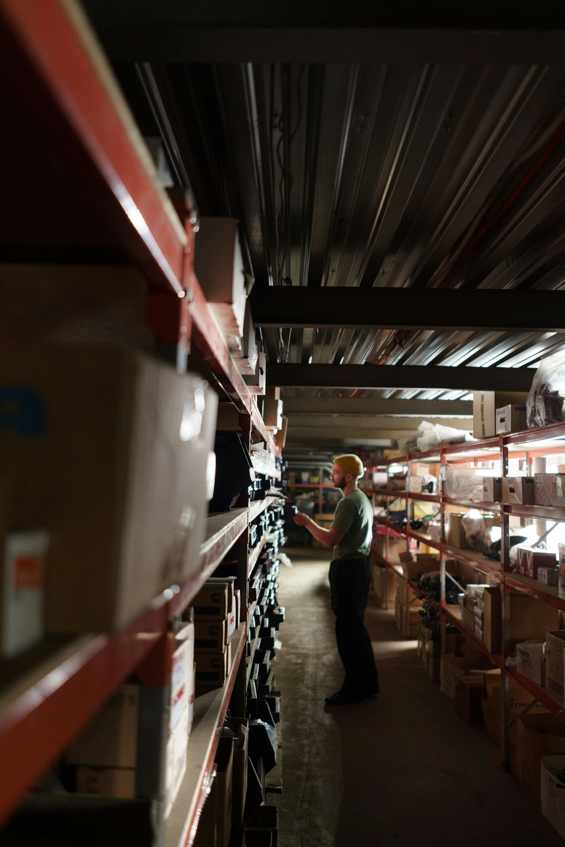 A person in a warehouse looking at boxes on shelves. Red shelving, dark room.