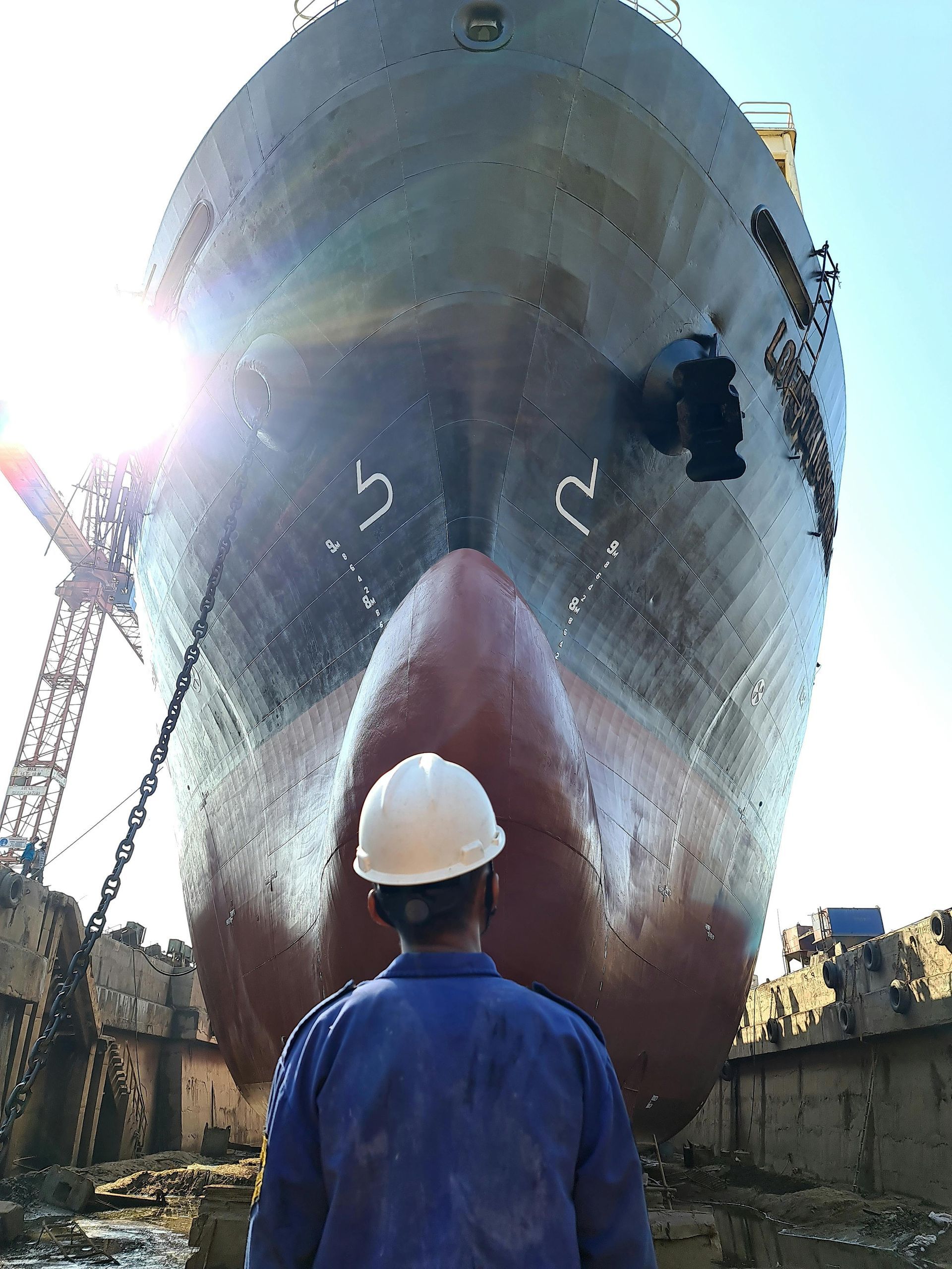 Worker in blue overalls and white hard hat looking up at the bow of a large ship in a drydock.