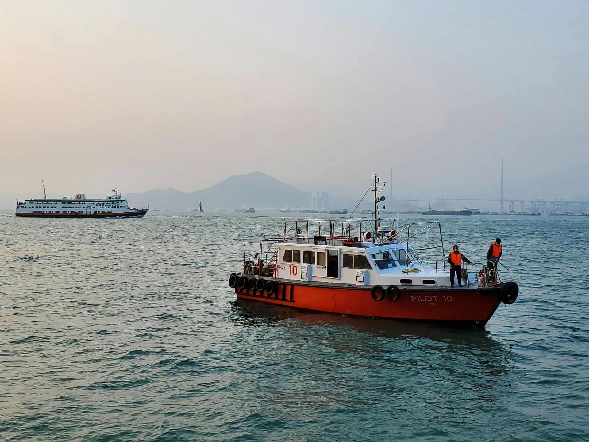 Orange and white boat with two people on board, in a body of water with other boats and a mountain in the background.