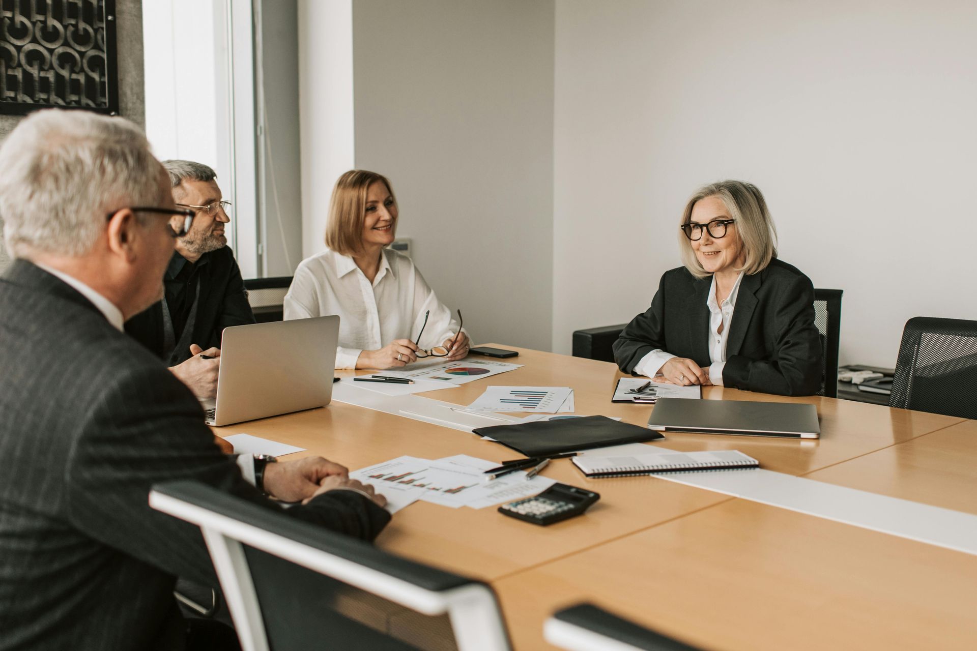Business meeting: Five people around a table, discussing documents and using laptops.