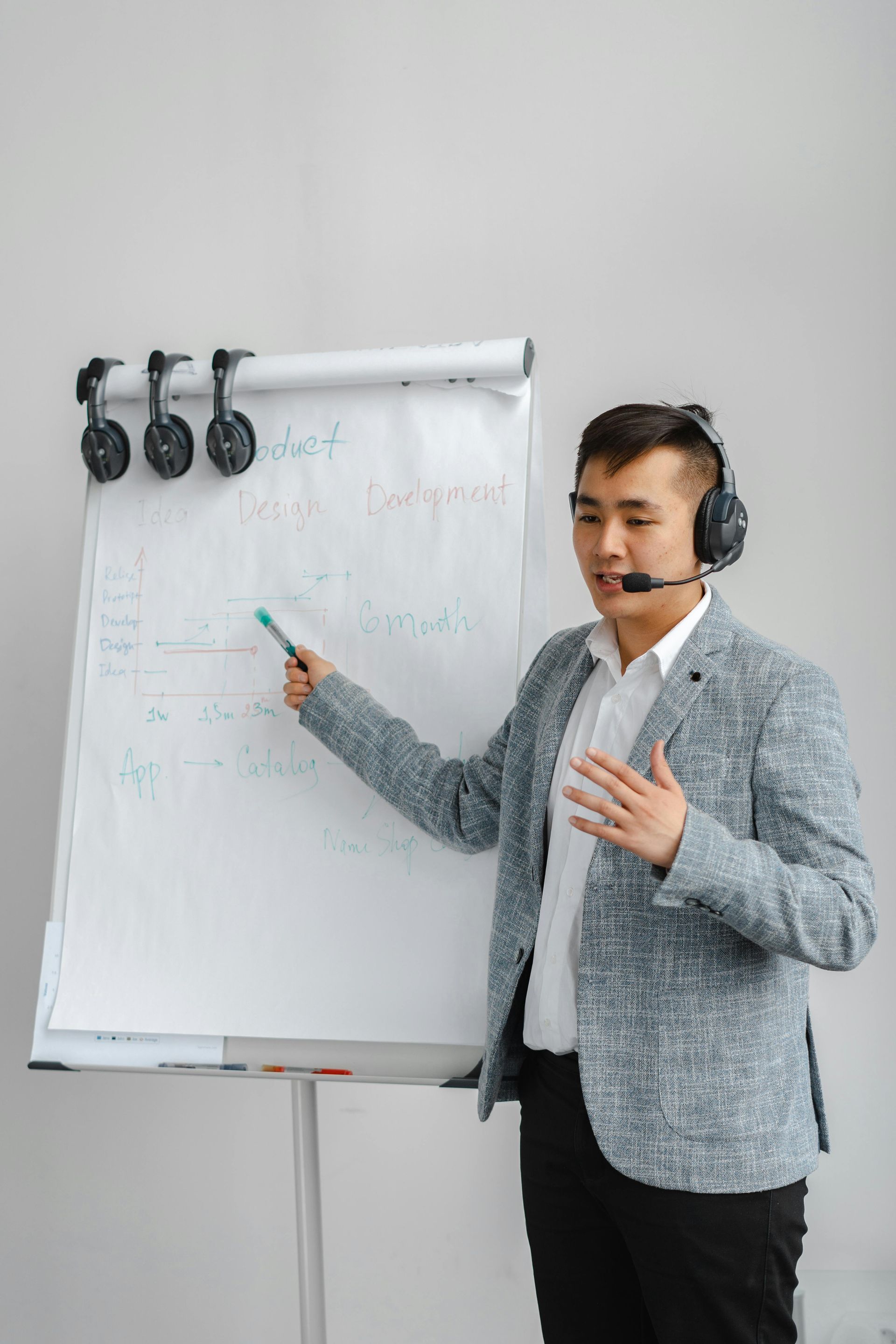 Man in blazer points to whiteboard, wearing headset; presentation setting.