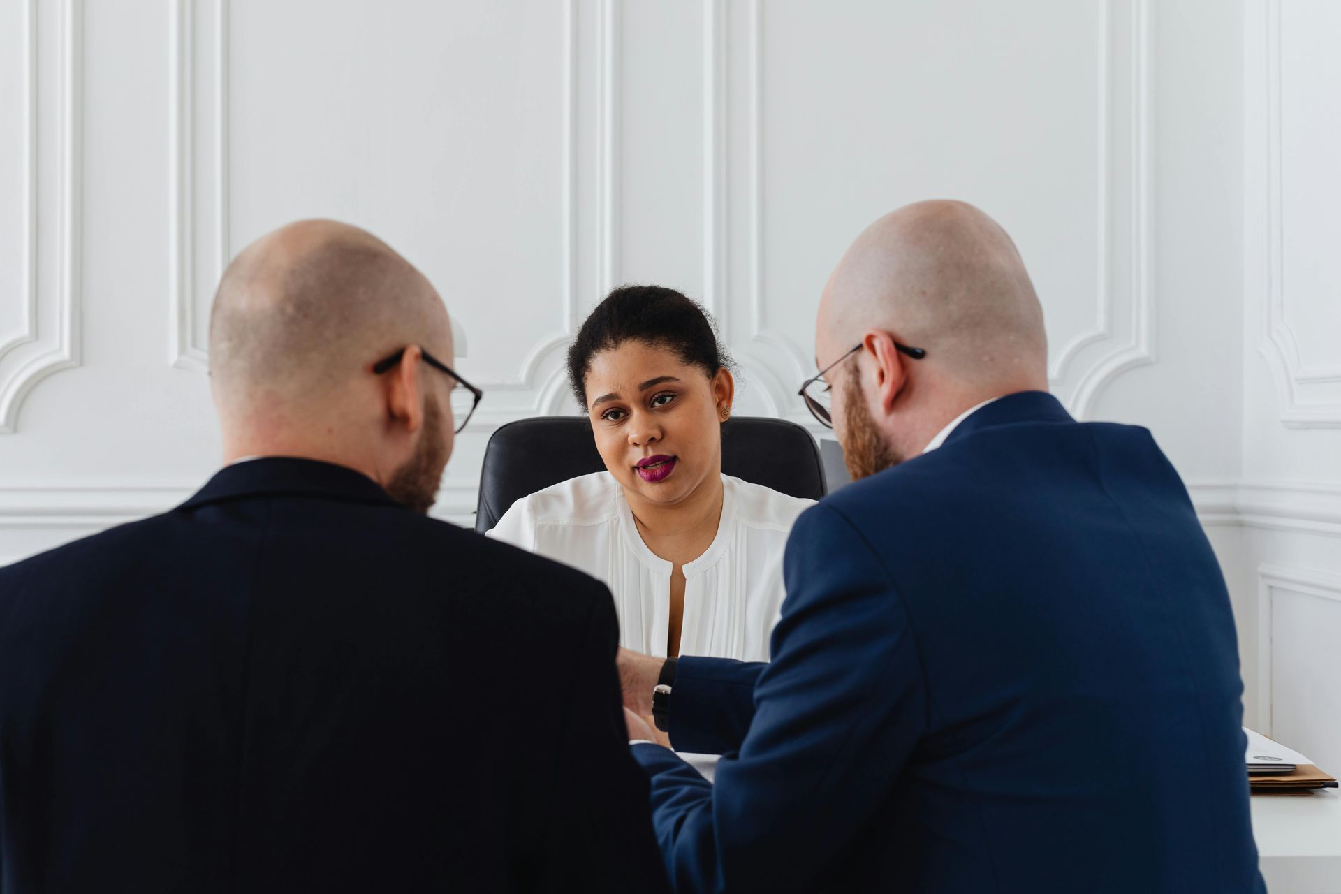 Two men in suits facing a woman at a desk; all are discussing in an office.