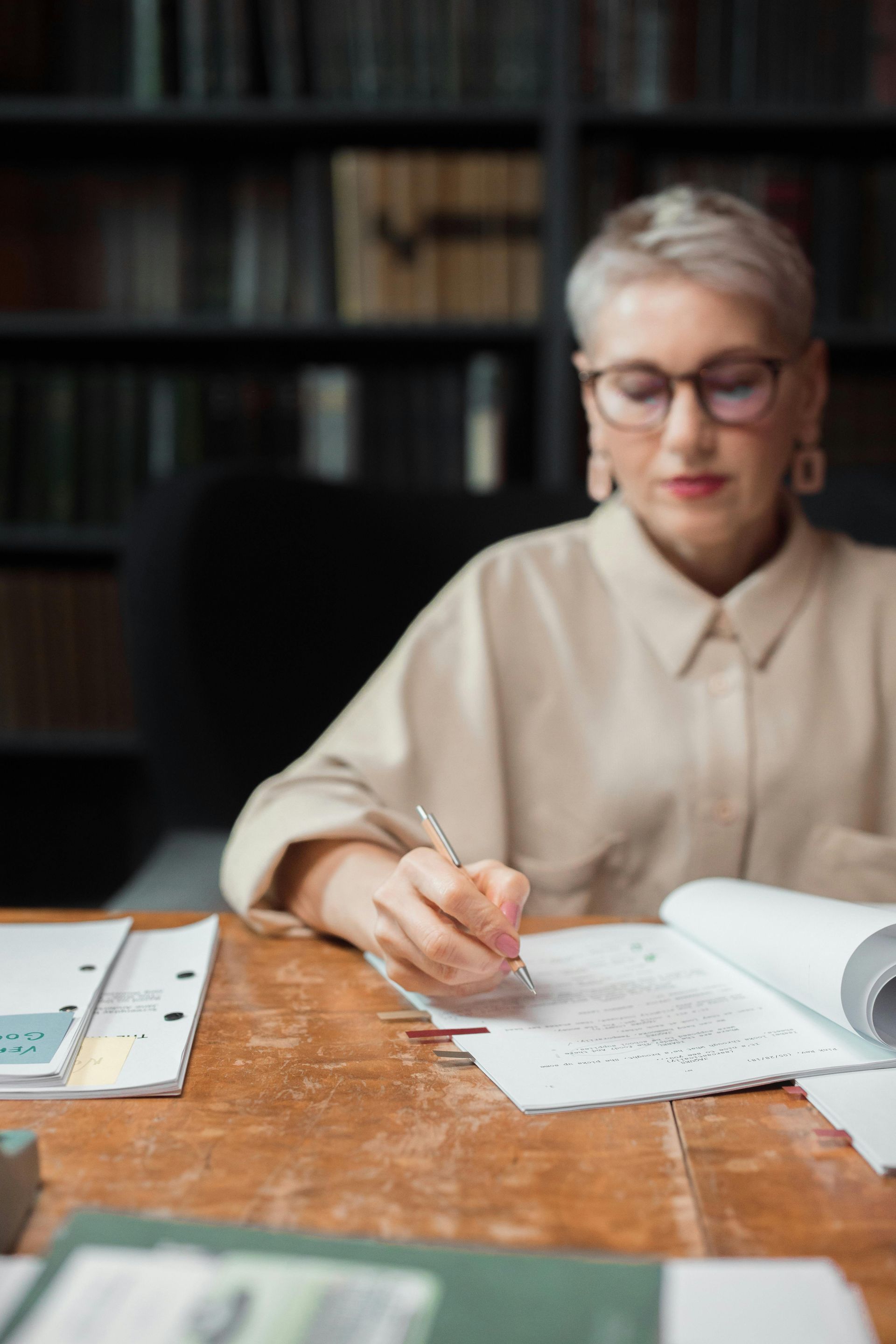 Woman with glasses and short hair writing at a wooden table, in a library.