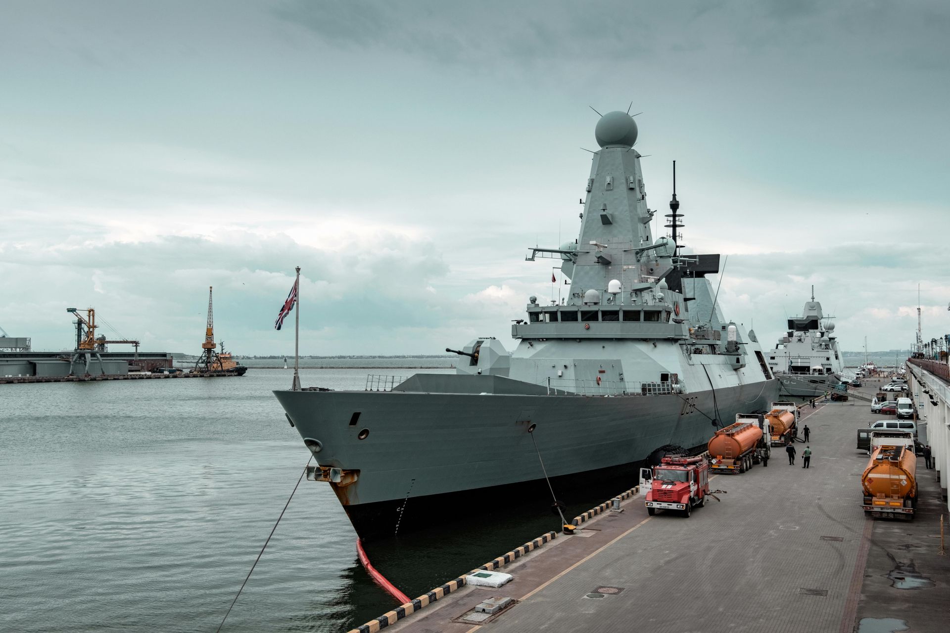 A gray Royal Navy warship docked in a harbor, with support vehicles and the Union Jack flag.