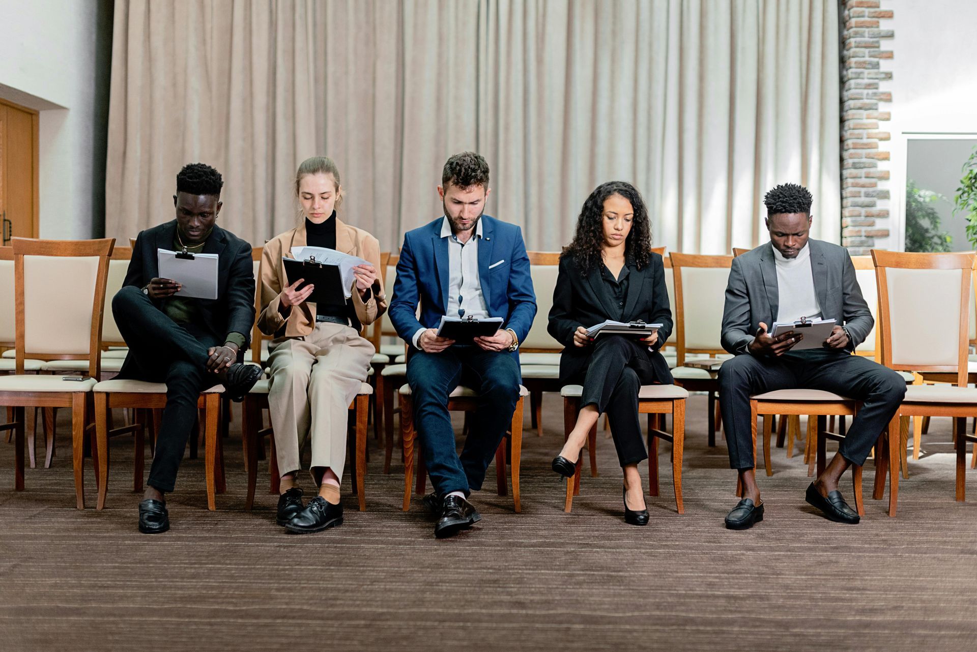 Five diverse people in business attire seated, holding papers, possibly awaiting interviews.