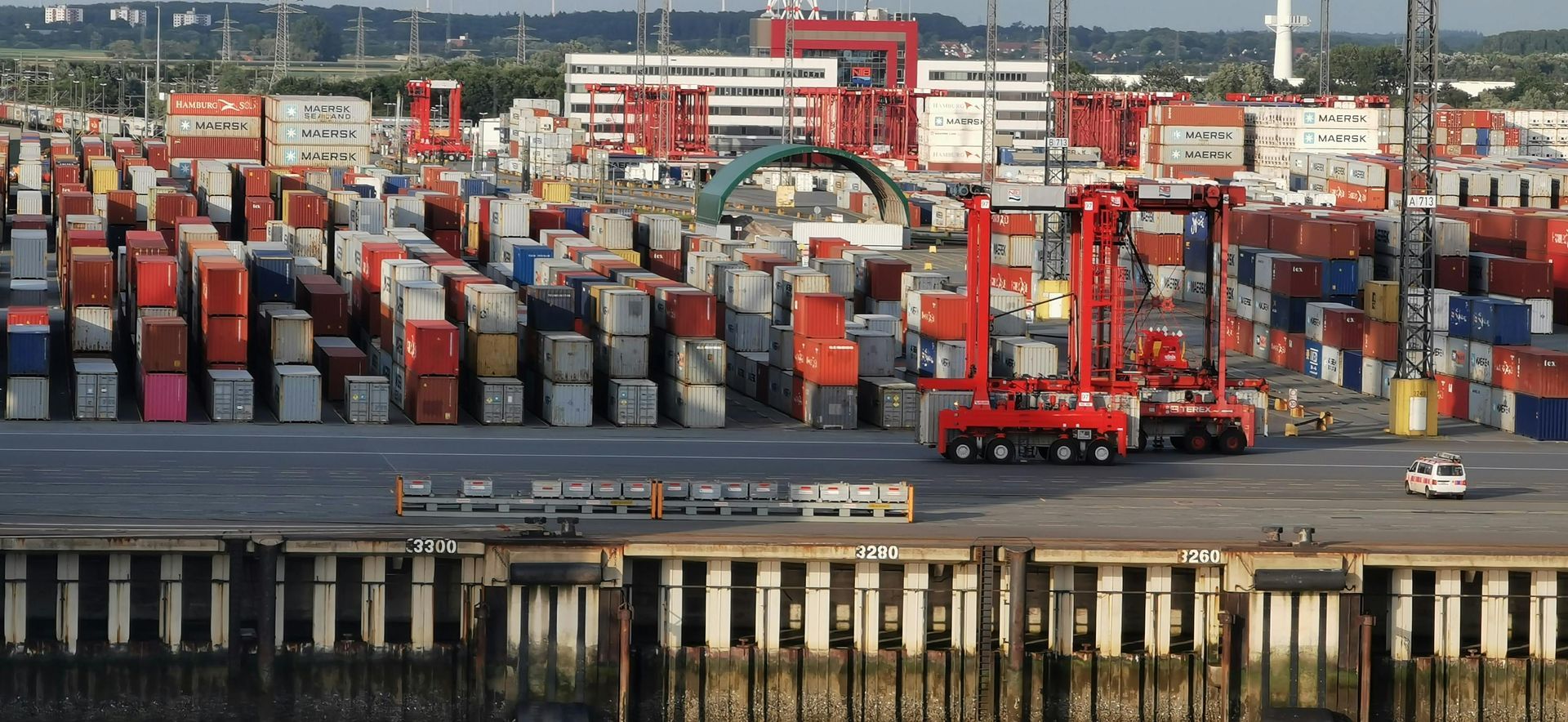 Shipping containers stacked in a port, with red cranes and a waterfront view.