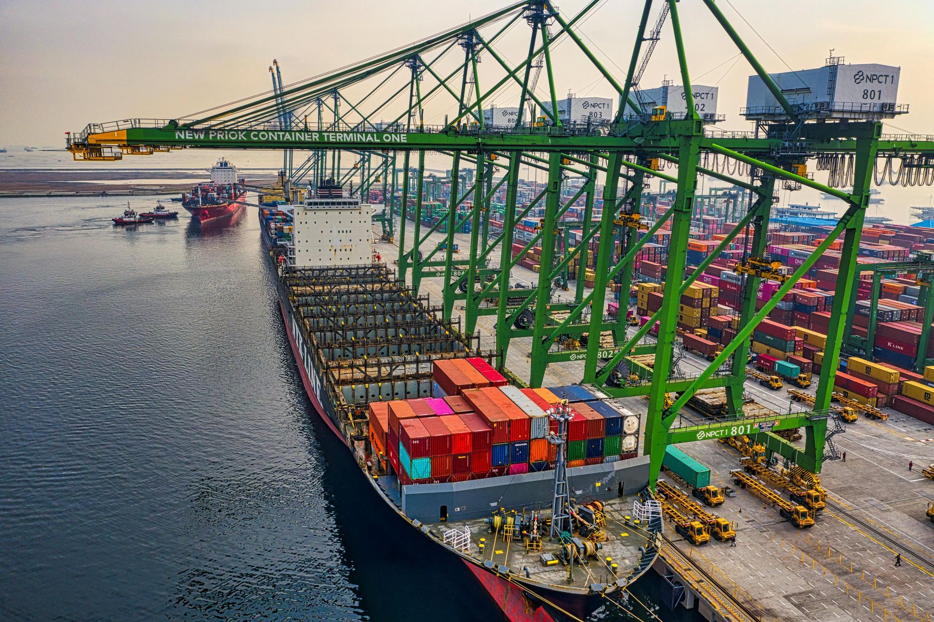 Cargo ship docked at a port with green cranes unloading containers; stacks of colorful shipping containers nearby.