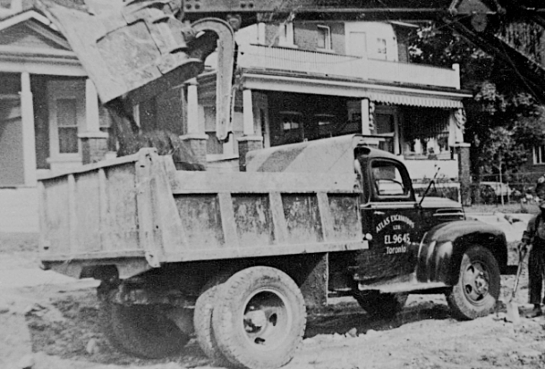 Photo en noir et blanc : Un camion à benne basculante est rempli de débris par une grue près d'un immeuble résidentiel.