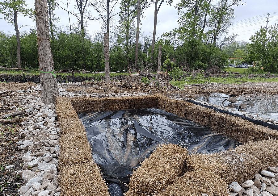 Construction site with dirt, pipes, and a pile of gravel, in a wooded area.