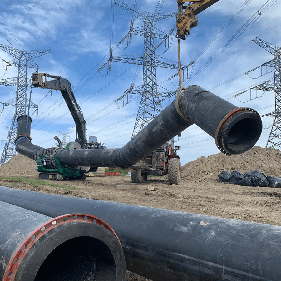 Construction workers using a large saw to cut a black pipeline outdoors; excavators in the background.