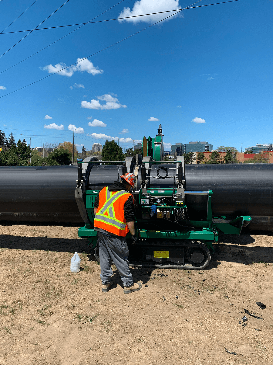 Construction workers using a machine to weld a large black pipe outdoors, with yellow equipment and a forest background.
