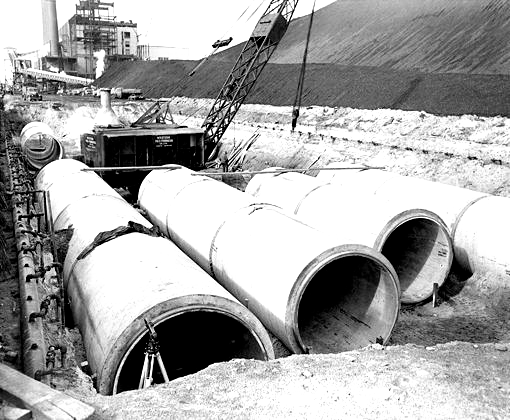 Gros tuyaux en béton dans une tranchée, avec une grue et un décor industriel en arrière-plan.