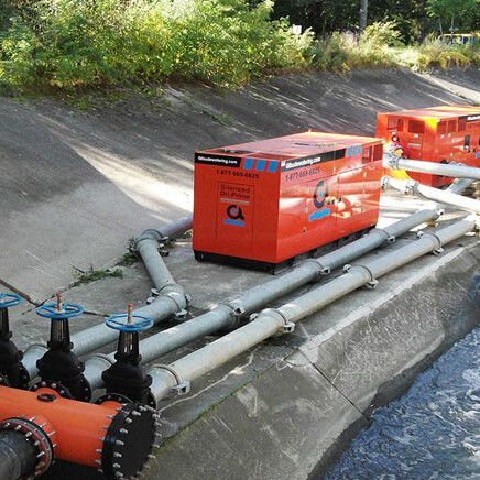 Des pompes à eau orange avec des tuyaux connectés sont placées le long d'un canal d'eau en béton.