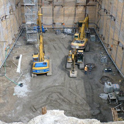 Fosse d'excavation avec trois véhicules de construction jaunes, entourée de murs en bois et d'échafaudages.