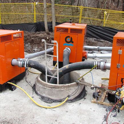 Three orange pumps connected to a manhole with hoses. A construction site with yellow fencing.