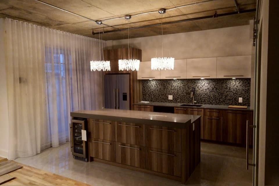 Cuisine moderne avec armoires en bois, îlot central et luminaires suspendus. Plafond et sol en béton apparent.